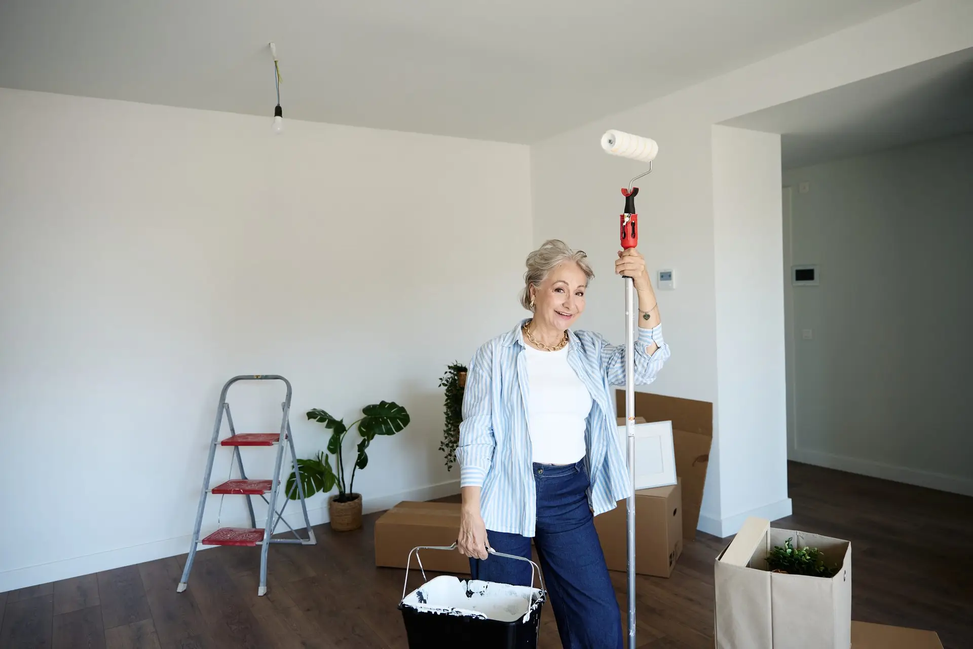 Femme tenant un rouleau télescopique et un bac de peinture dans une pièce vide aux murs blancs, escabeau et cartons